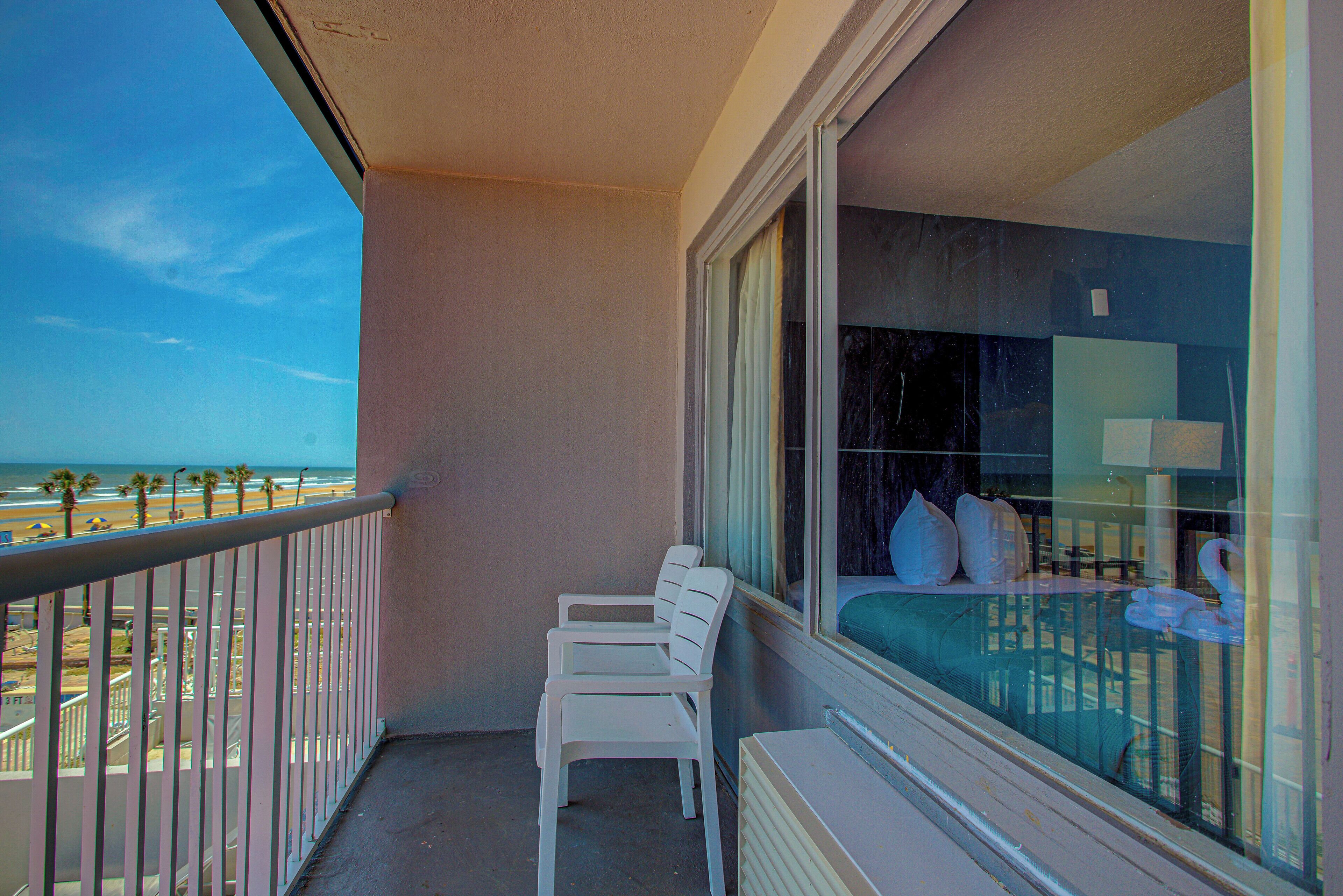 A balcony with a view of the beach, a bed with pillows, and a chair.