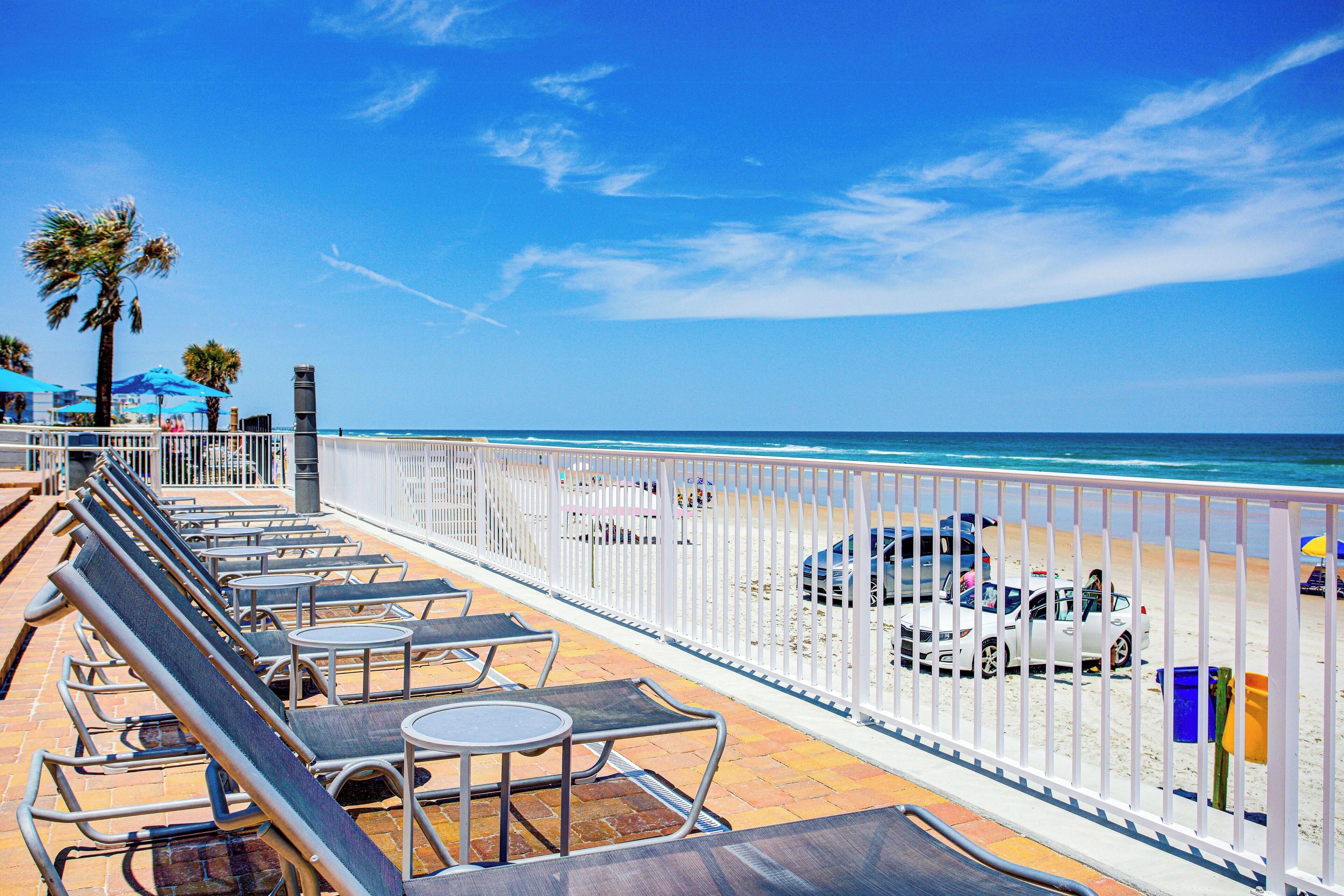 A beachside lounge with sun loungers and tables, a white fence, and a view of the ocean.