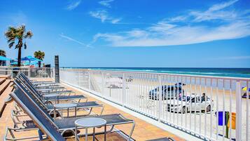 A beachside lounge with sun loungers and tables, a white fence, and a view of the ocean.