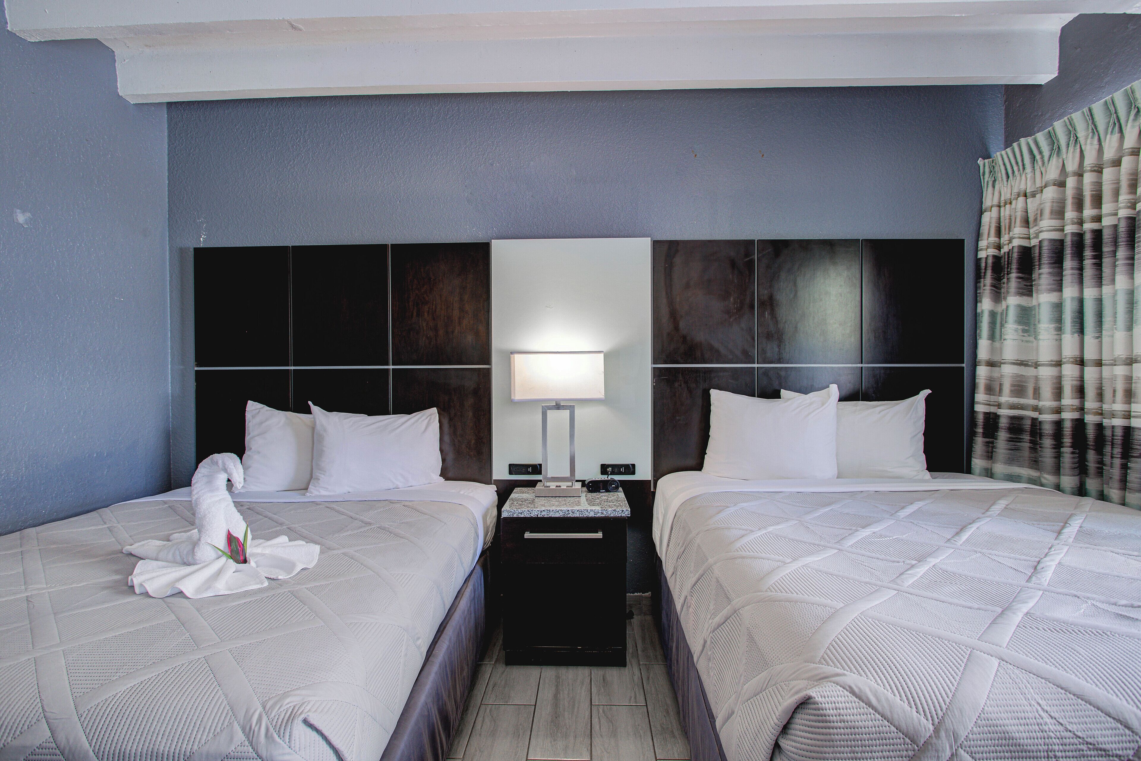 A hotel room with two beds, a nightstand, a lamp, and a wall-mounted mirror.
