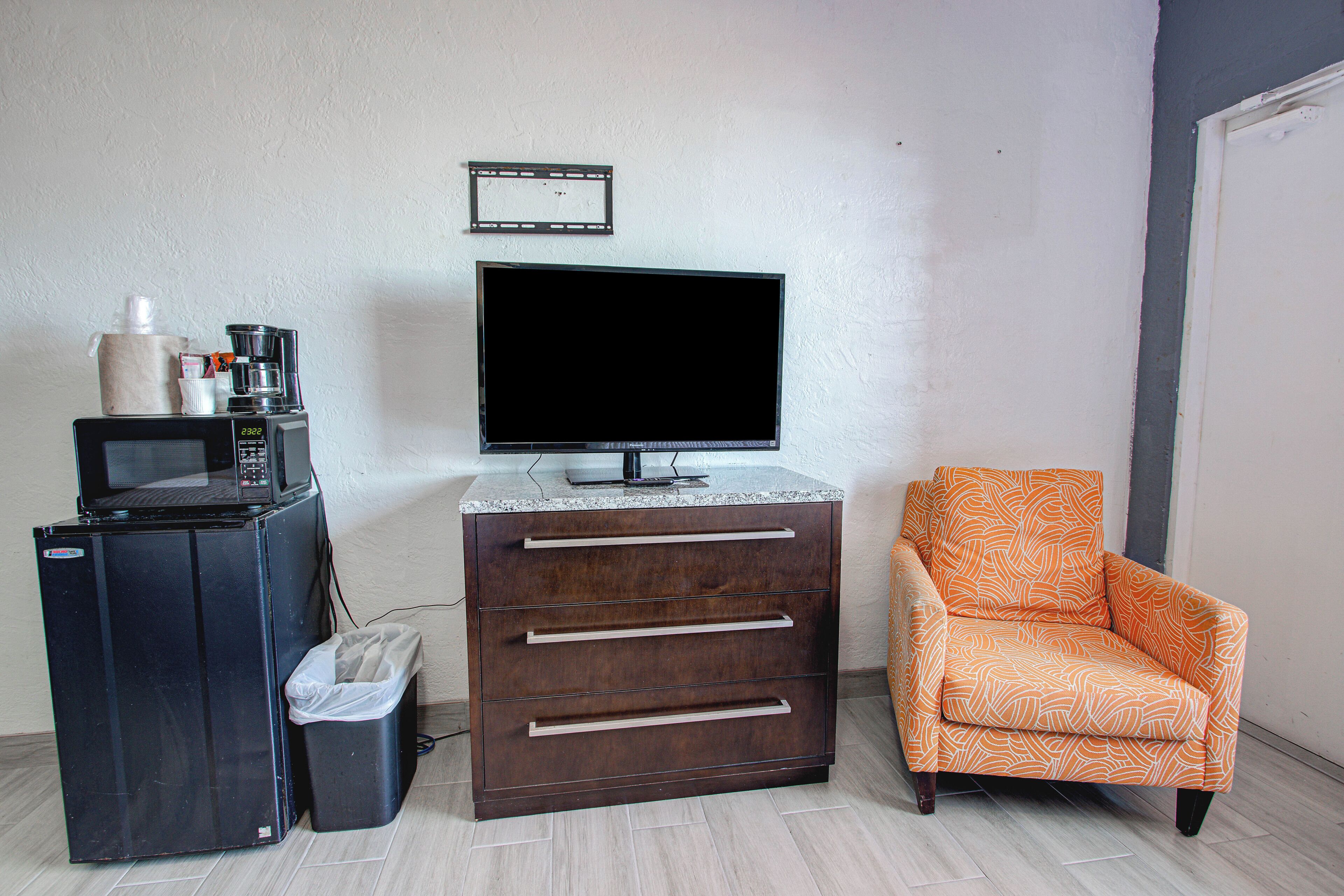 A room with a television, a coffee maker, a trash bin, a cabinet with drawers, and an orange patterned chair.