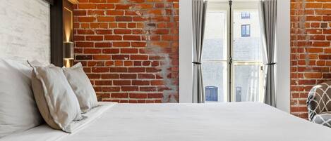 A hotel room with a brick wall, a bed with white linens, a nightstand with a lamp, and a view through a glass door.