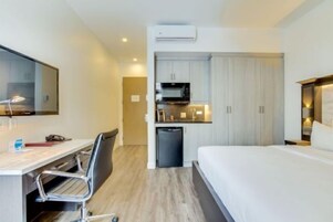 Habitación de hotel con cama, escritorio y silla, kitchenette con microondas y refrigerador pequeño.