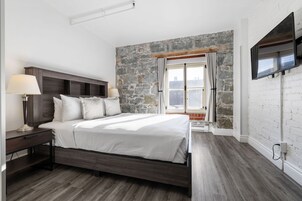 Un dormitorio con pared de piedra, una cama con ropa de cama blanca, una mesita de noche con lámpara y un televisor montado en la pared.