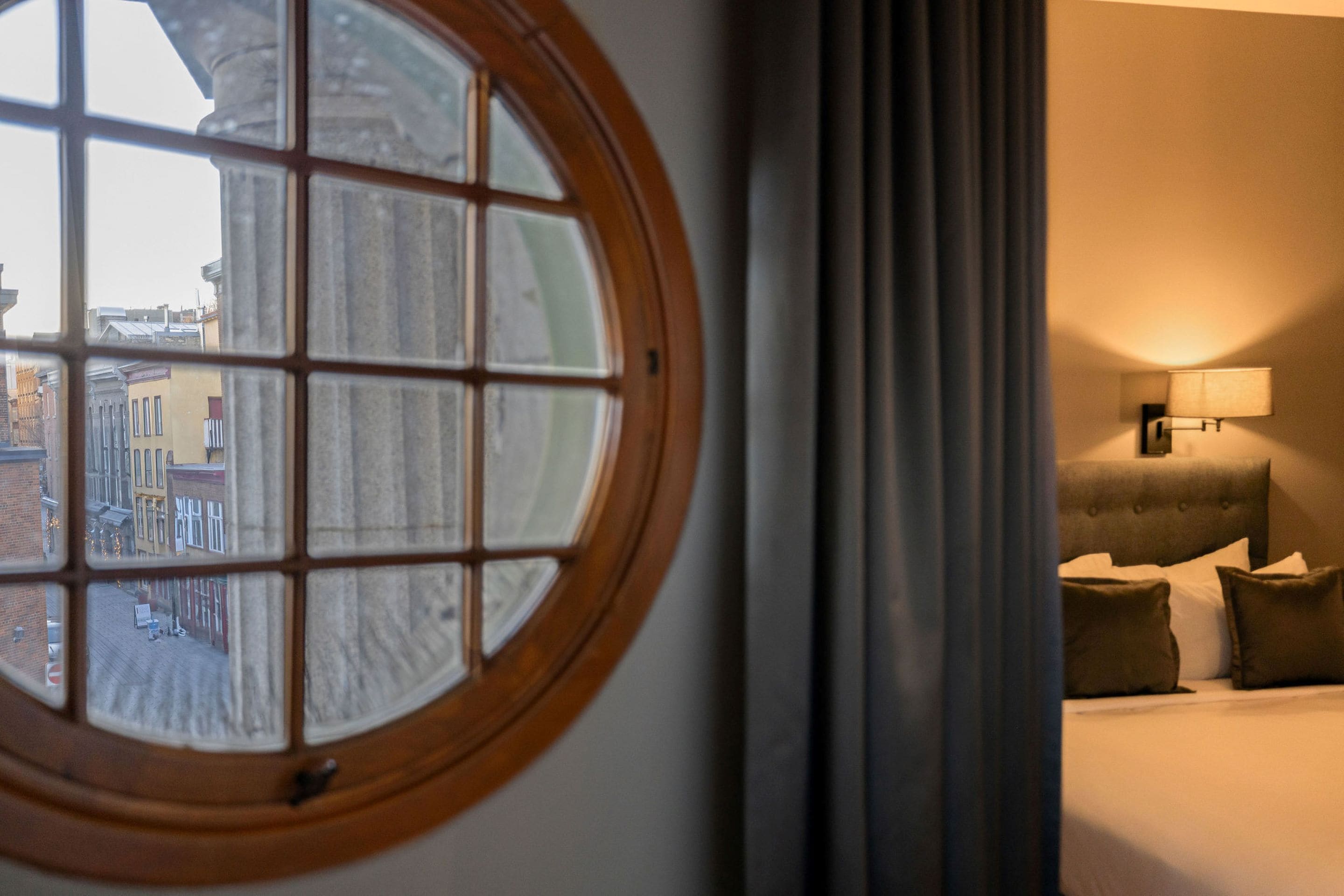 A hotel room with a large window, a bed with pillows, and a wall-mounted lamp.