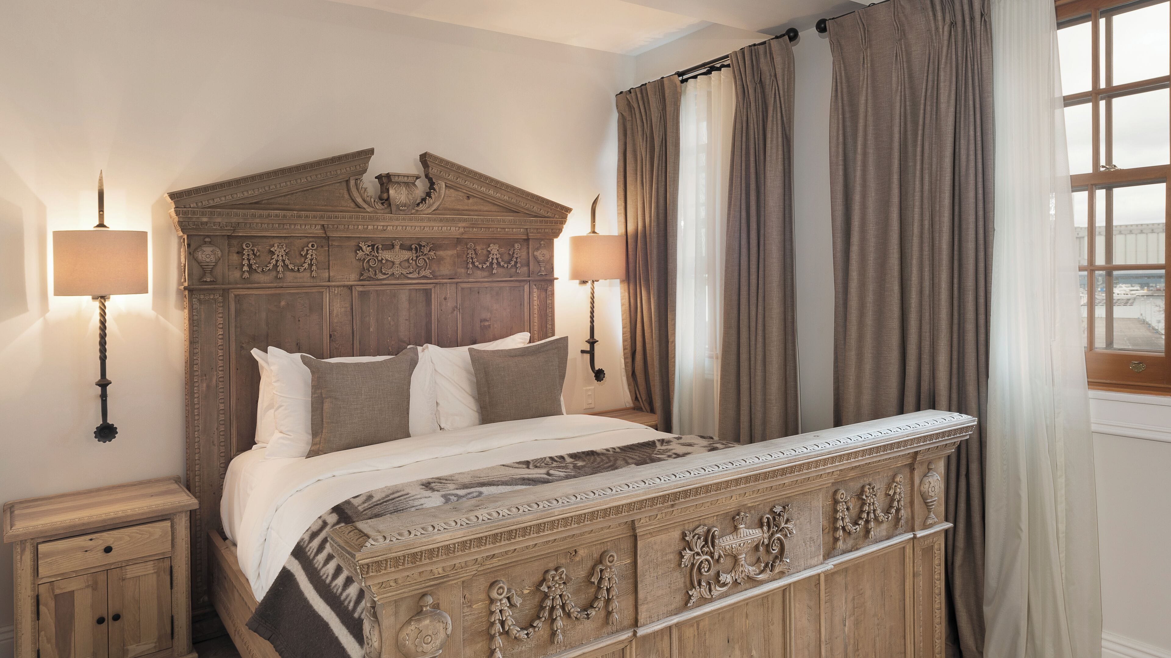 Een klassiek houten bed met decoratieve houtsnijwerken, twee bedlampen en uitzicht op het groen door een raam met gordijnen.