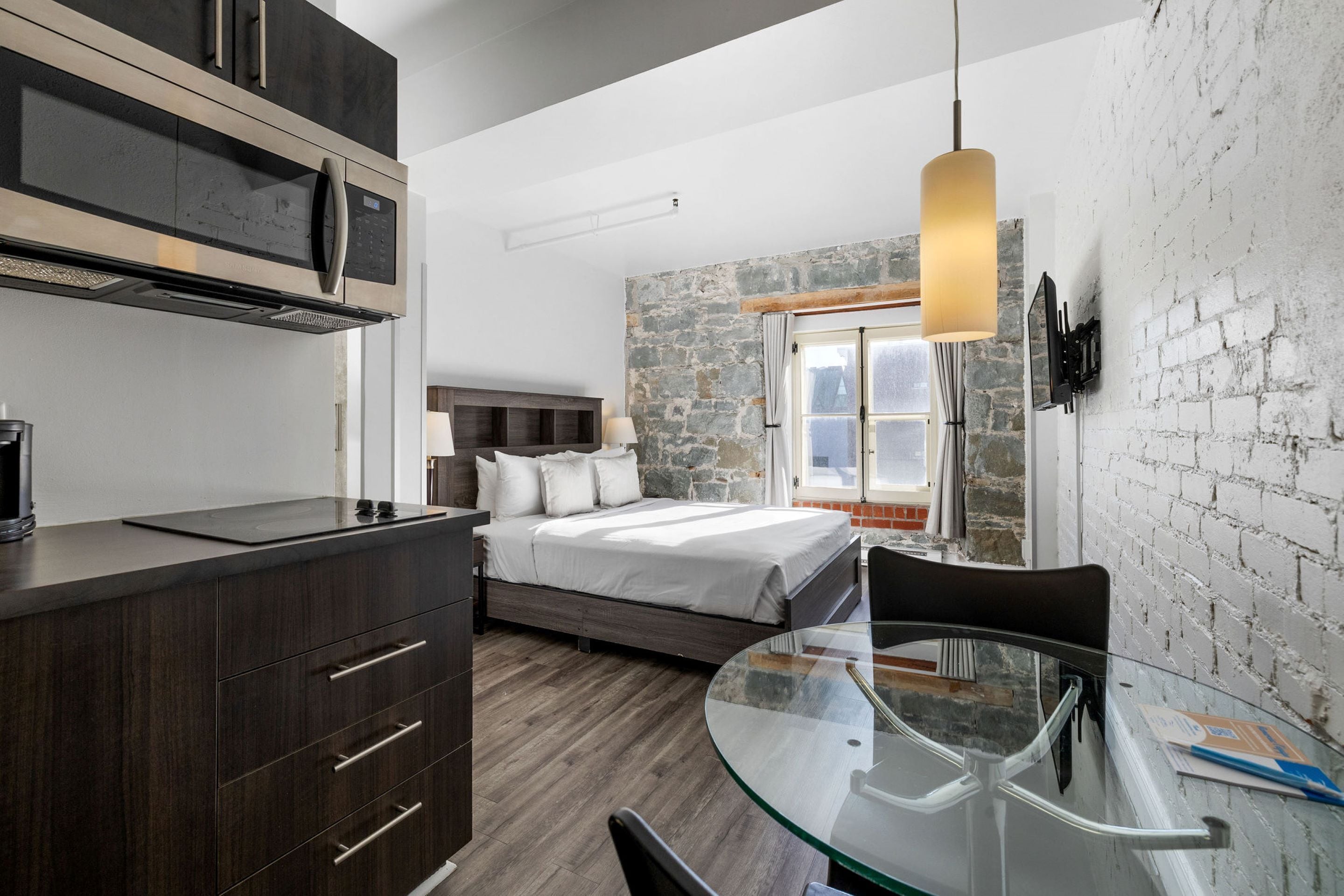 Une chambre d’hôtel compacte comprenant un lit, une table à manger en verre, un coin cuisine et un mur en pierre.