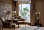 A hotel room with a large window, a potted plant, a chair, a table with a lamp, and a bed.