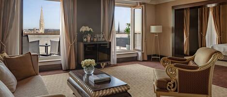 Ein Hotelzimmer mit großem Fenster und Blick auf eine Stadtansicht, darunter ein markanter Turm. Das Zimmer ist mit einer Couch, einem Sessel, einem kleinen Tisch mit Blumenvase und einer Lampe ausgestattet.