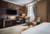 A hotel room with a large flat-screen TV mounted on the wall, a desk with a coffee machine, a chair, a round table with a bowl of fruit, and a bed.