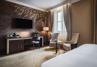 A hotel room with a large flat-screen TV mounted on the wall, a desk with a coffee machine, a chair, a round table with a bowl of fruit, and a bed.