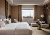 A modern hotel room with a large bed, two armchairs, a small table, and a view of a cityscape.