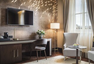 A modern hotel room with a large flat-screen TV, a desk with a coffee maker, a chair, a lamp, and a view of the city through the window.