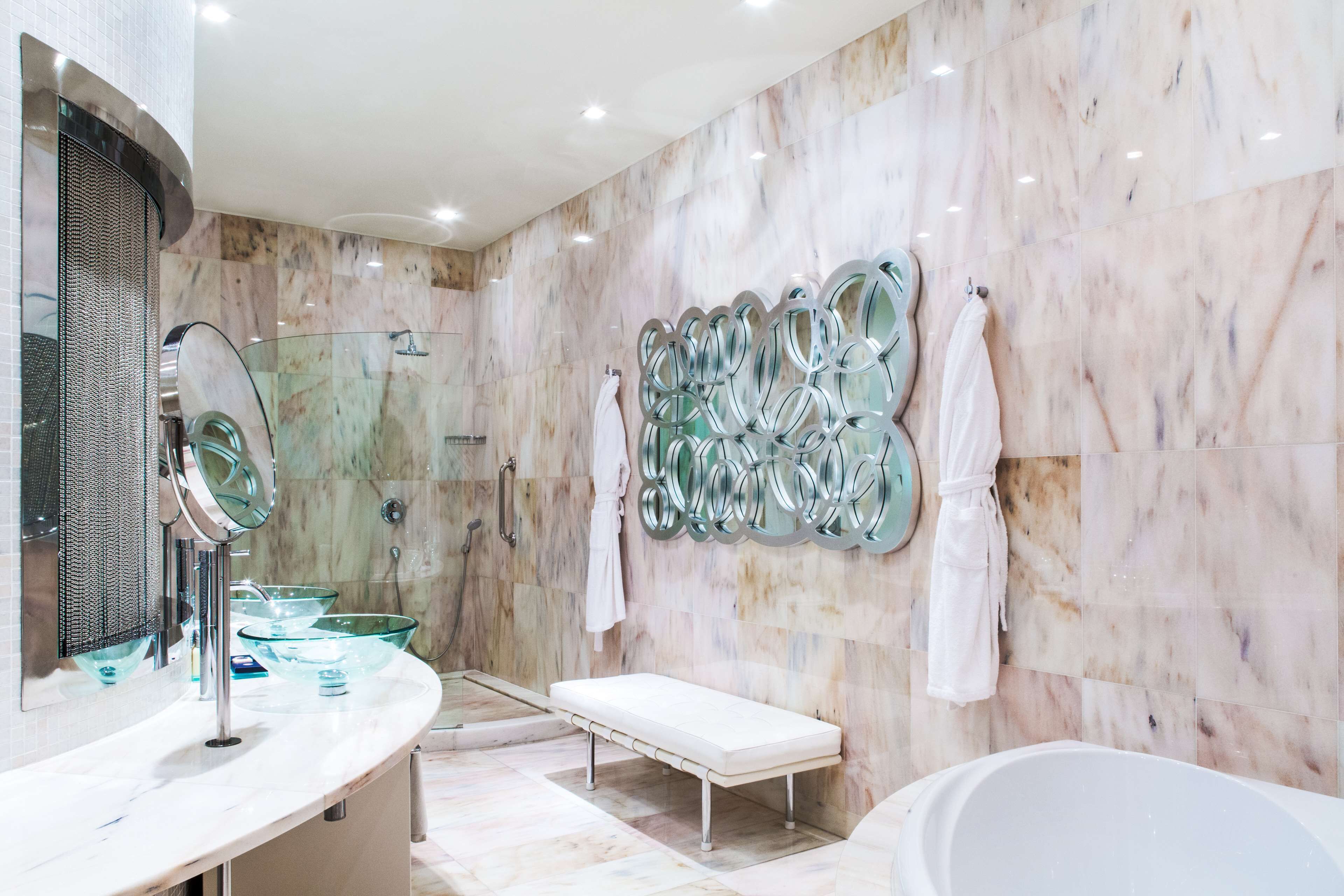A modern bathroom with a marble wall, a glass shower enclosure, a white bench, and a bathtub.