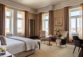 A hotel room with a large bed, two armchairs, a small table, and a view of the city.