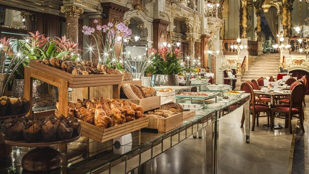 Un lobby de hotel lujoso con arquitectura ornamentada, una larga mesa de buffet con diversos pasteles y elegantes candelabros.