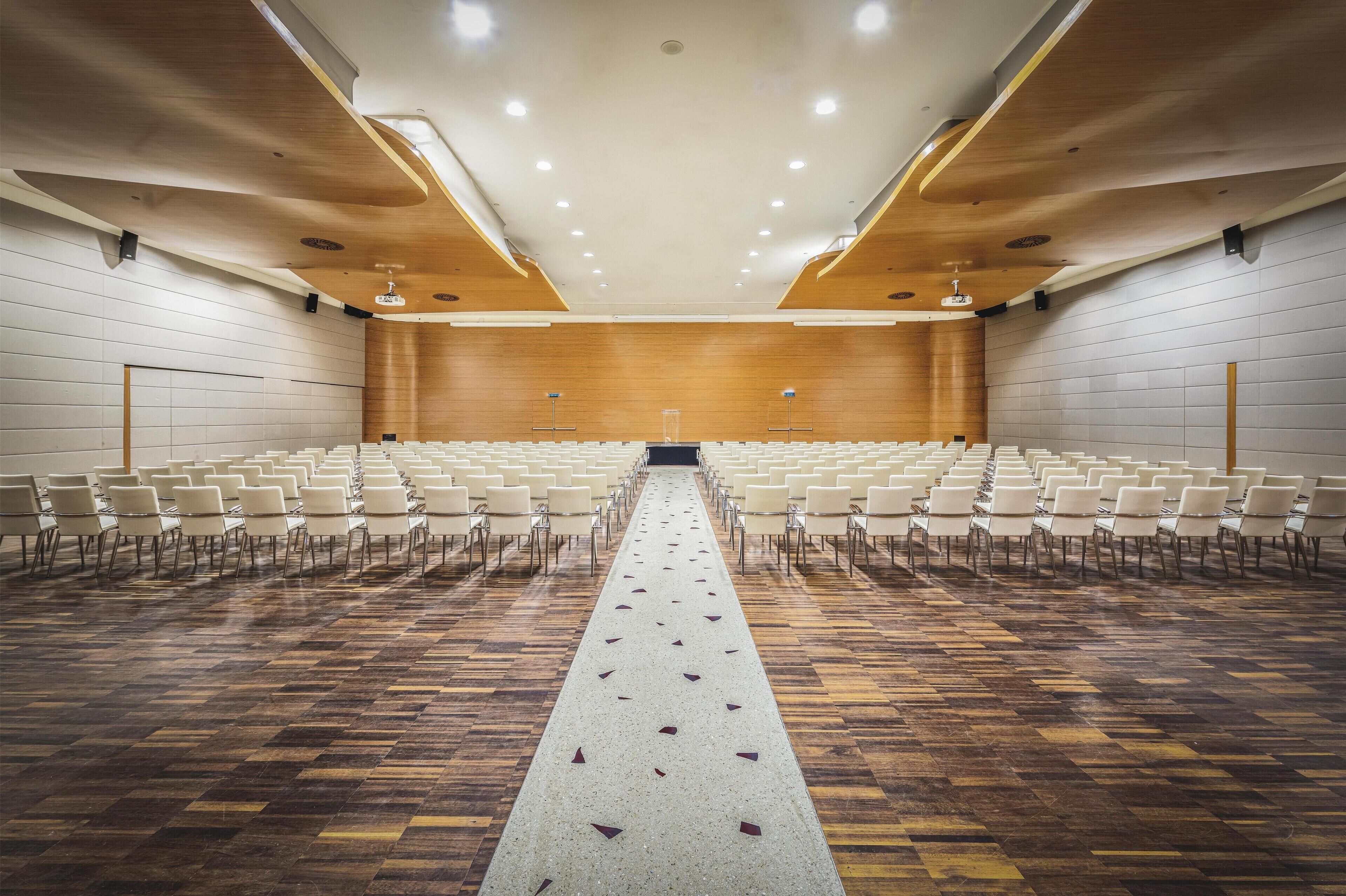 Un salón de conferencias con filas de sillas, un pasillo central y suelo de madera.