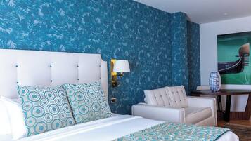 A hotel room with a bed, a white headboard, patterned pillows, a blue patterned wall, a table with a vase, and a chair.