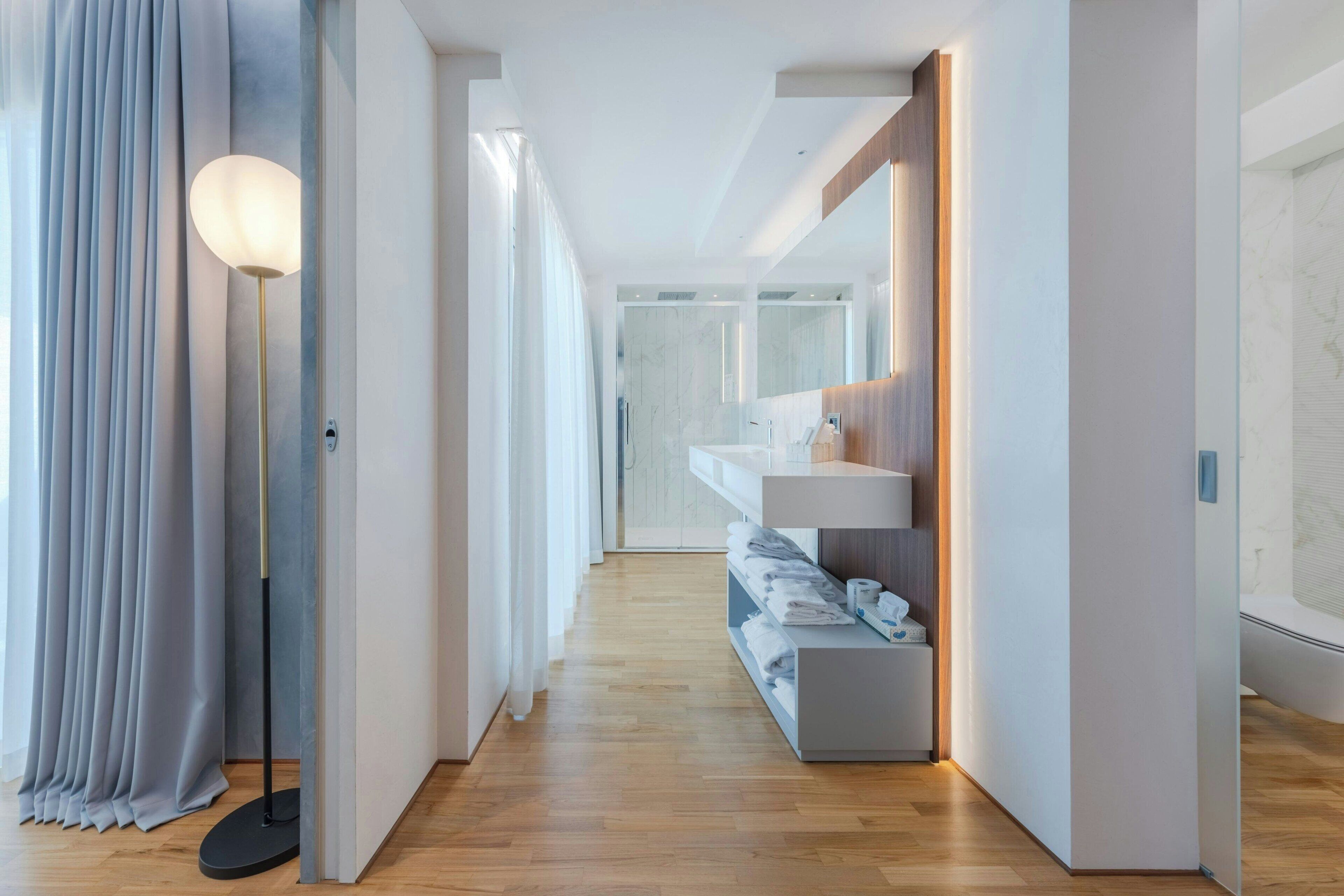 A modern hotel corridor with a wooden floor, a white reception desk, and a large mirror reflecting the interior.