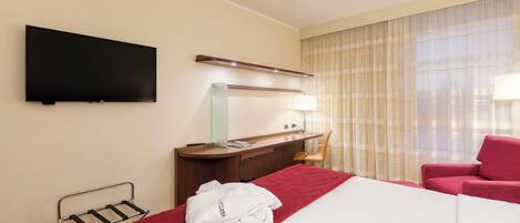Una habitación de hotel con cama, escritorio, silla, televisor y ventana con cortinas.