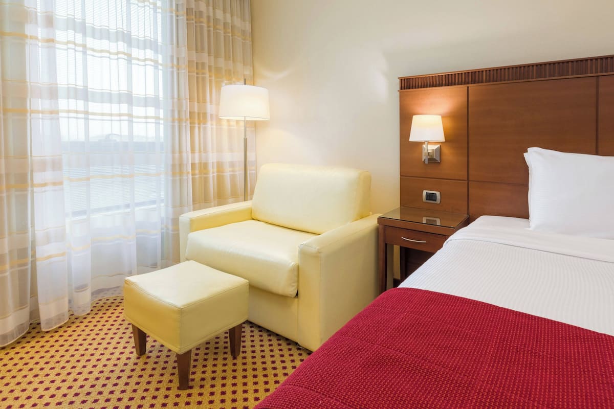 Une chambre d’hôtel comprenant un lit, une chaise, un pouf, une table de chevet, une lampe et une fenêtre avec des rideaux légers.
