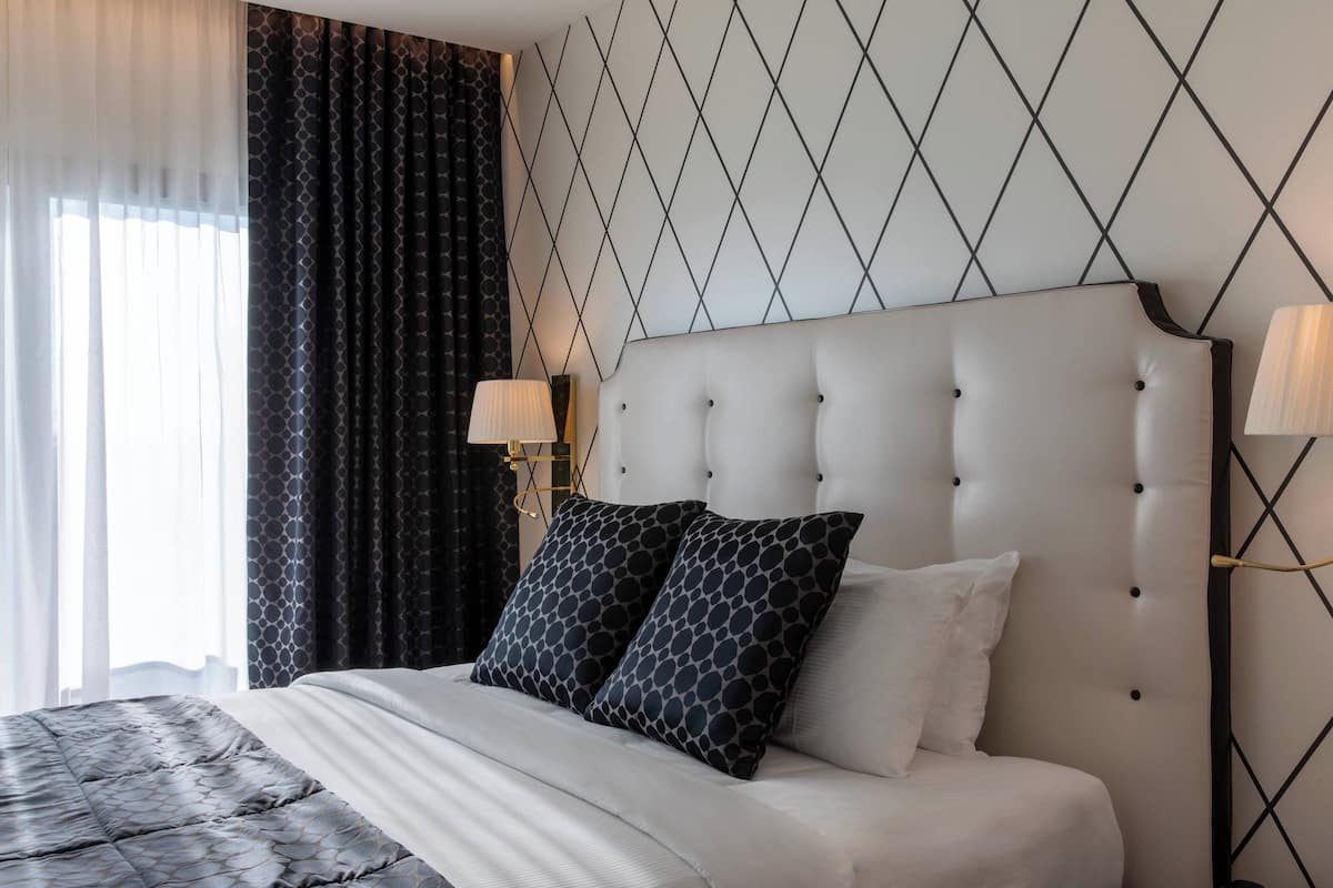 Une chambre d’hôtel avec un grand lit, une tête de lit capitonnée, deux lampes fixées au mur et un rideau à motifs géométriques.