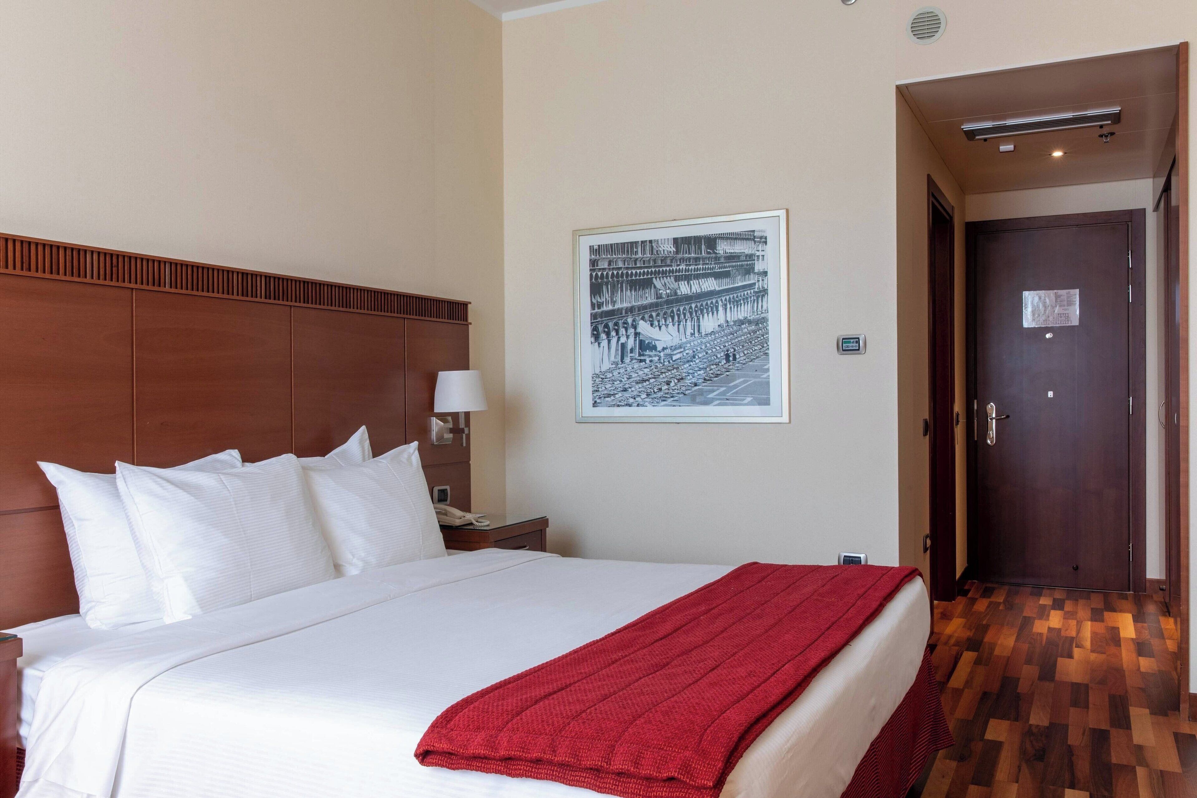 A hotel room with a large bed, a wooden headboard, a nightstand with a lamp, a framed picture on the wall, and a door leading to another room.
