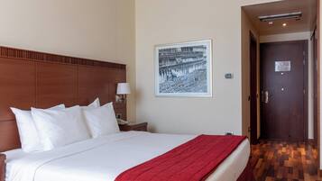 A hotel room with a large bed, a wooden headboard, a nightstand with a lamp, a framed picture on the wall, and a door leading to another room.