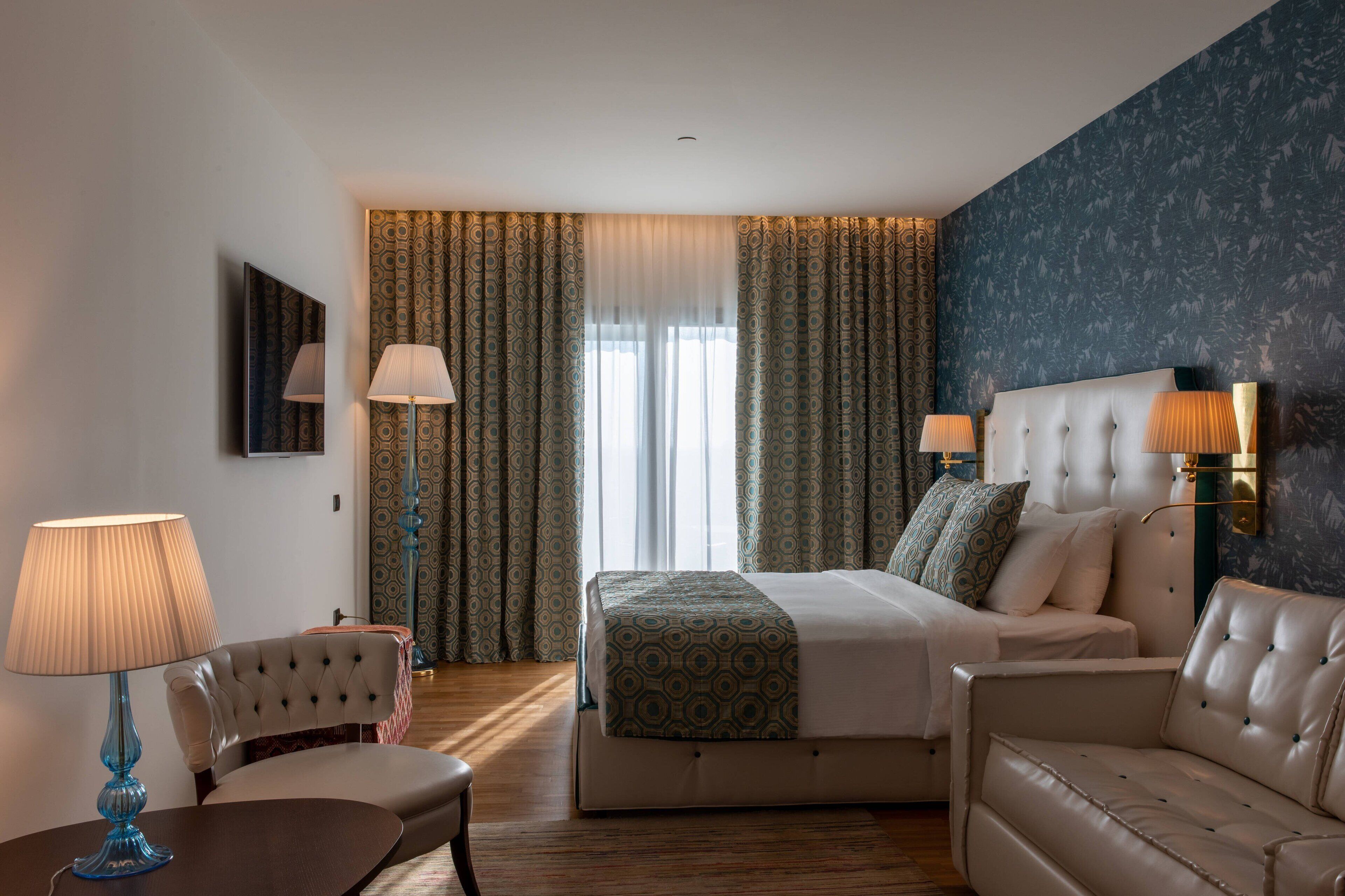 A hotel room with a large bed, two bedside lamps, a sofa, a chair, and a TV on the wall.