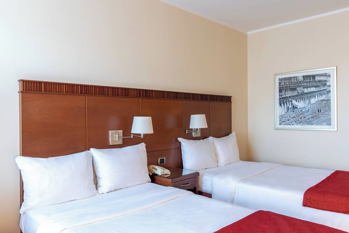 Une chambre d’hôtel avec deux lits, une tête de lit en bois, des lampes de chevet, un téléphone et un tableau encadré au mur.