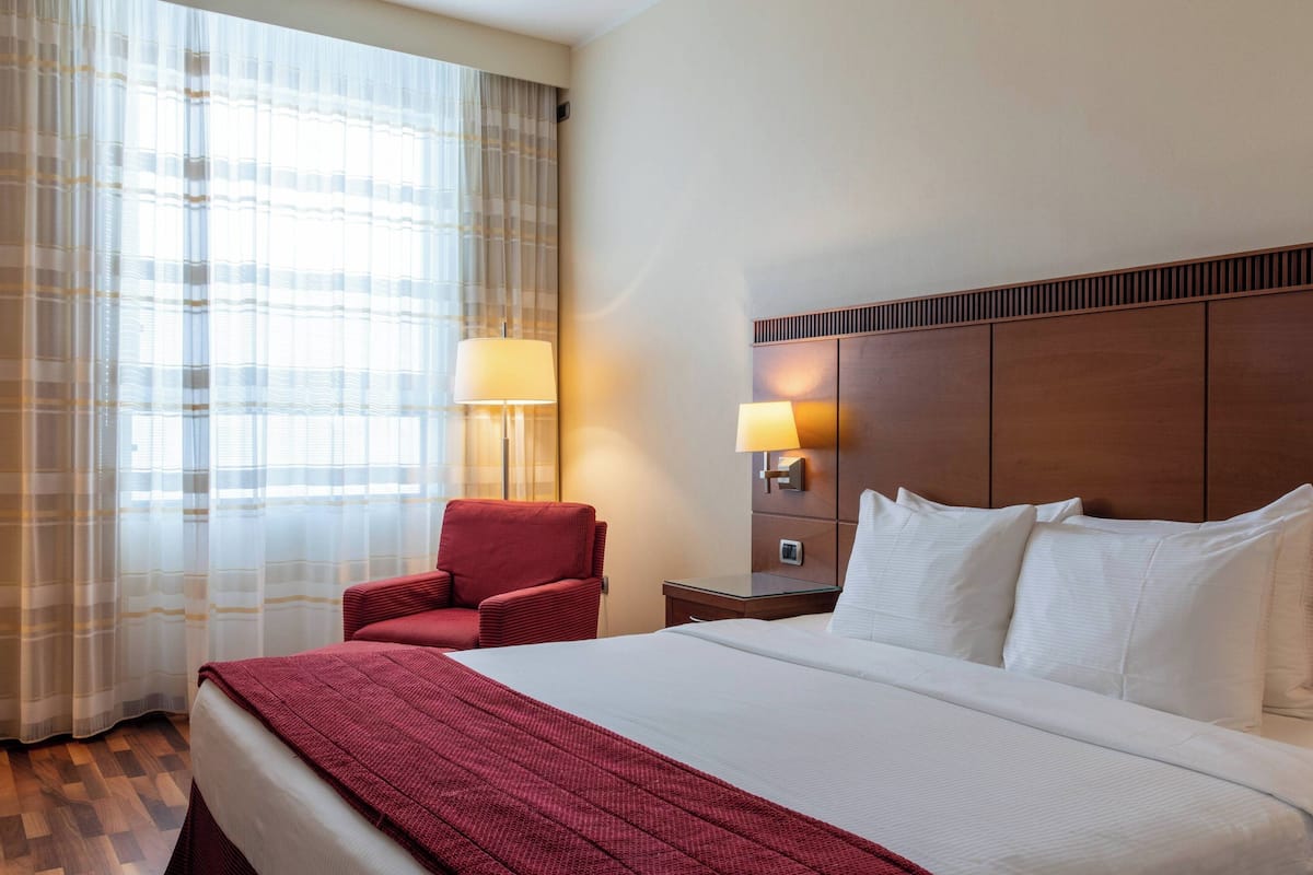 Une chambre d’hôtel avec un grand lit, un fauteuil rouge, une table de chevet avec une lampe et une fenêtre avec des rideaux.