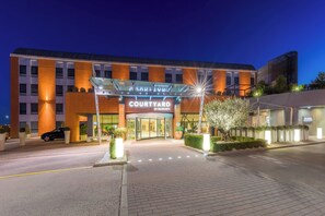 L’entrée d’un hôtel Courtyard by Marriott au crépuscule, avec un design moderne, une façade en verre et un chemin bien éclairé.