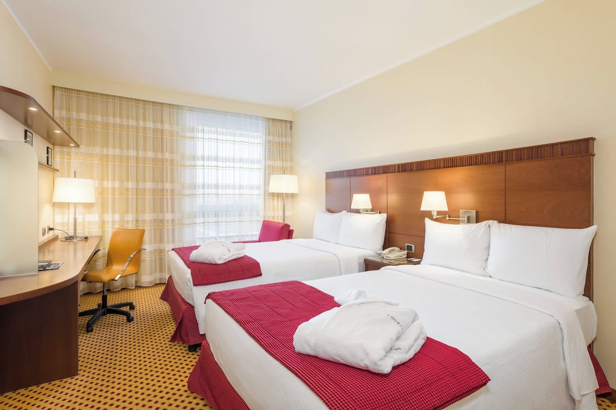 Une chambre d’hôtel avec deux lits, un bureau et une chaise. Il y a des lampes sur les tables de chevet et une fenêtre avec des rideaux.