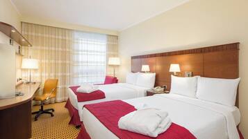 A hotel room with two beds, a desk, and a chair. There are lamps on the nightstands and a window with curtains.