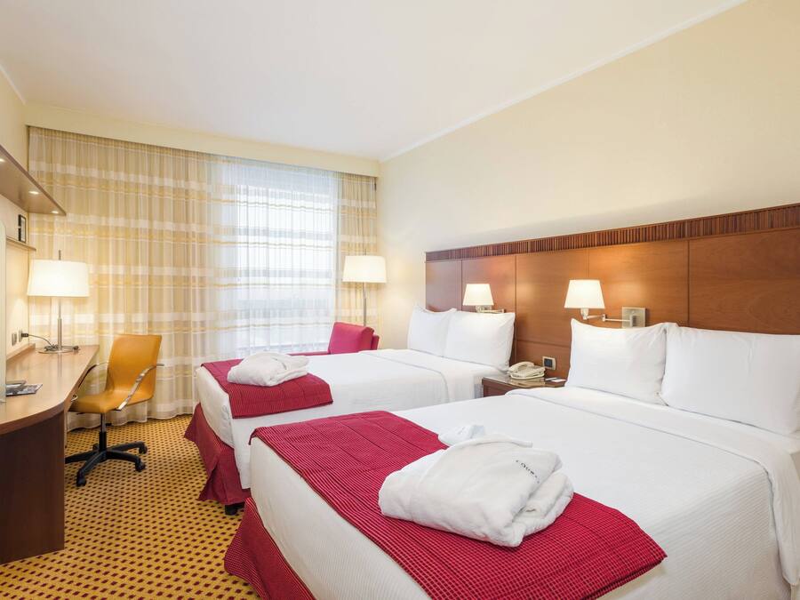 A hotel room with two beds, a desk, and a chair. There are lamps on the nightstands and a window with curtains.