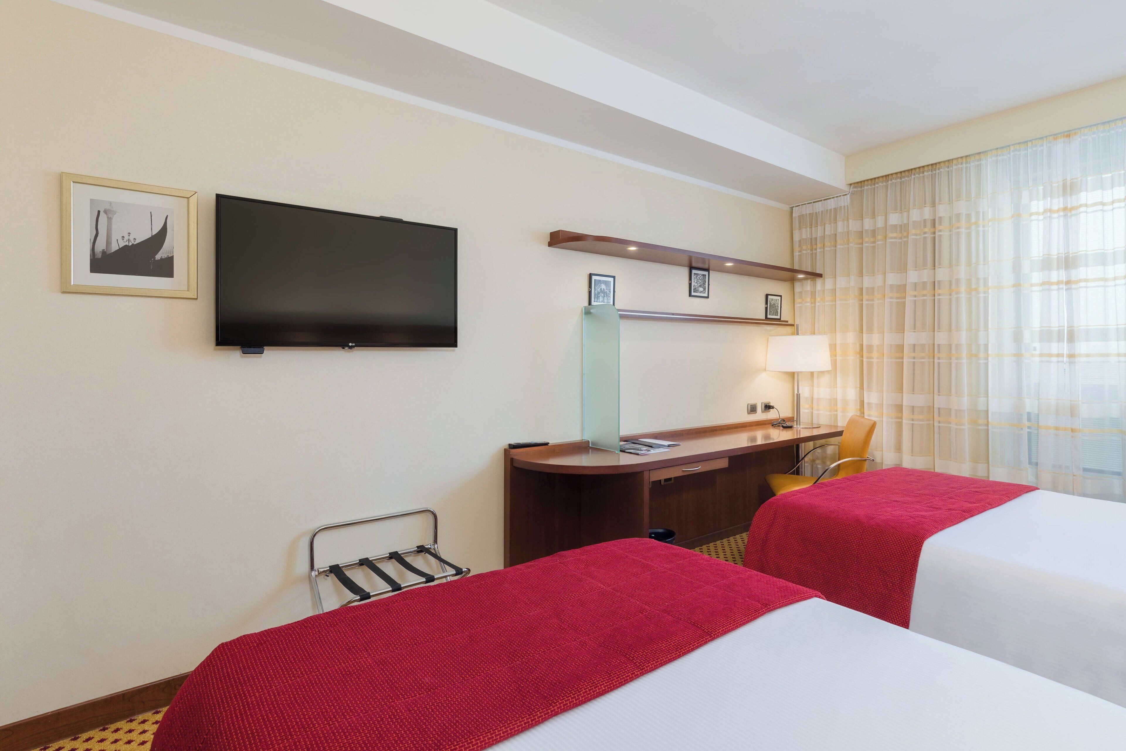Una habitación de hotel con dos camas, un escritorio, una silla, un televisor y un ventanal con cortinas.