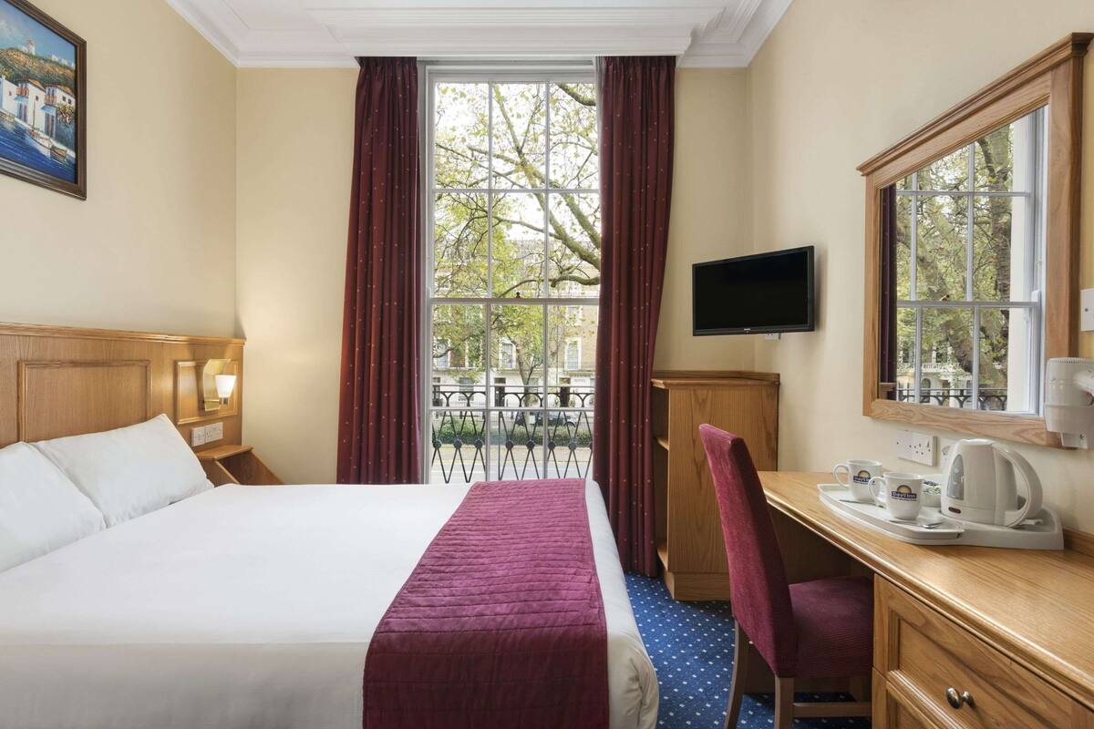 A hotel room with a bed, a desk with a chair, a television, and a window with curtains.