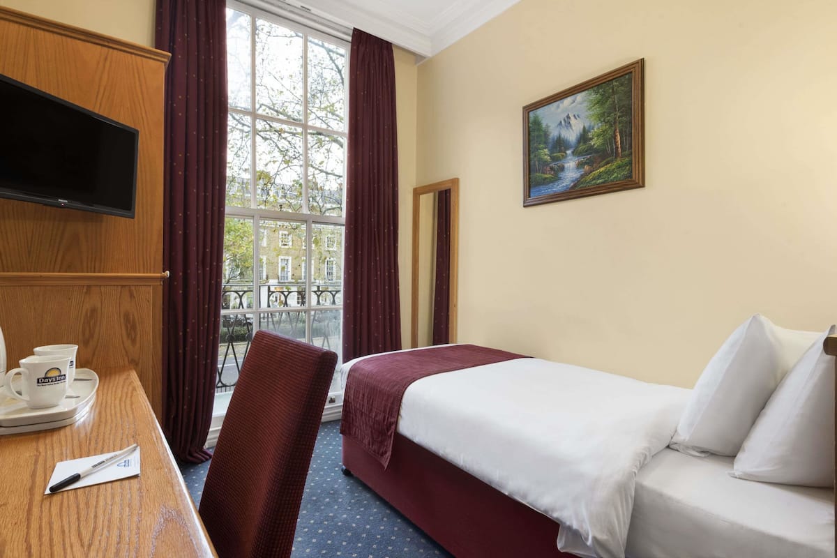 A hotel room with a bed, a TV, a painting, a window with curtains, and a table with a cup and saucer.
