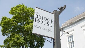Letrero de 'The Bridge House Bed & Breakfast' con vista despejada de un edificio blanco y un árbol grande al fondo.