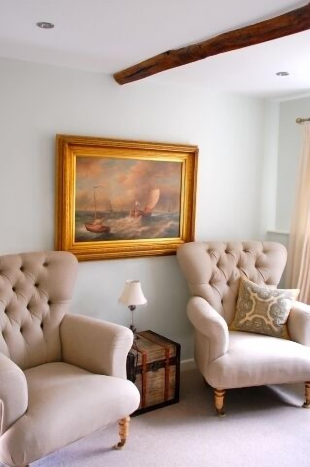 A room with two upholstered armchairs, a framed painting, a small table with a lamp, and a wooden beam on the ceiling.