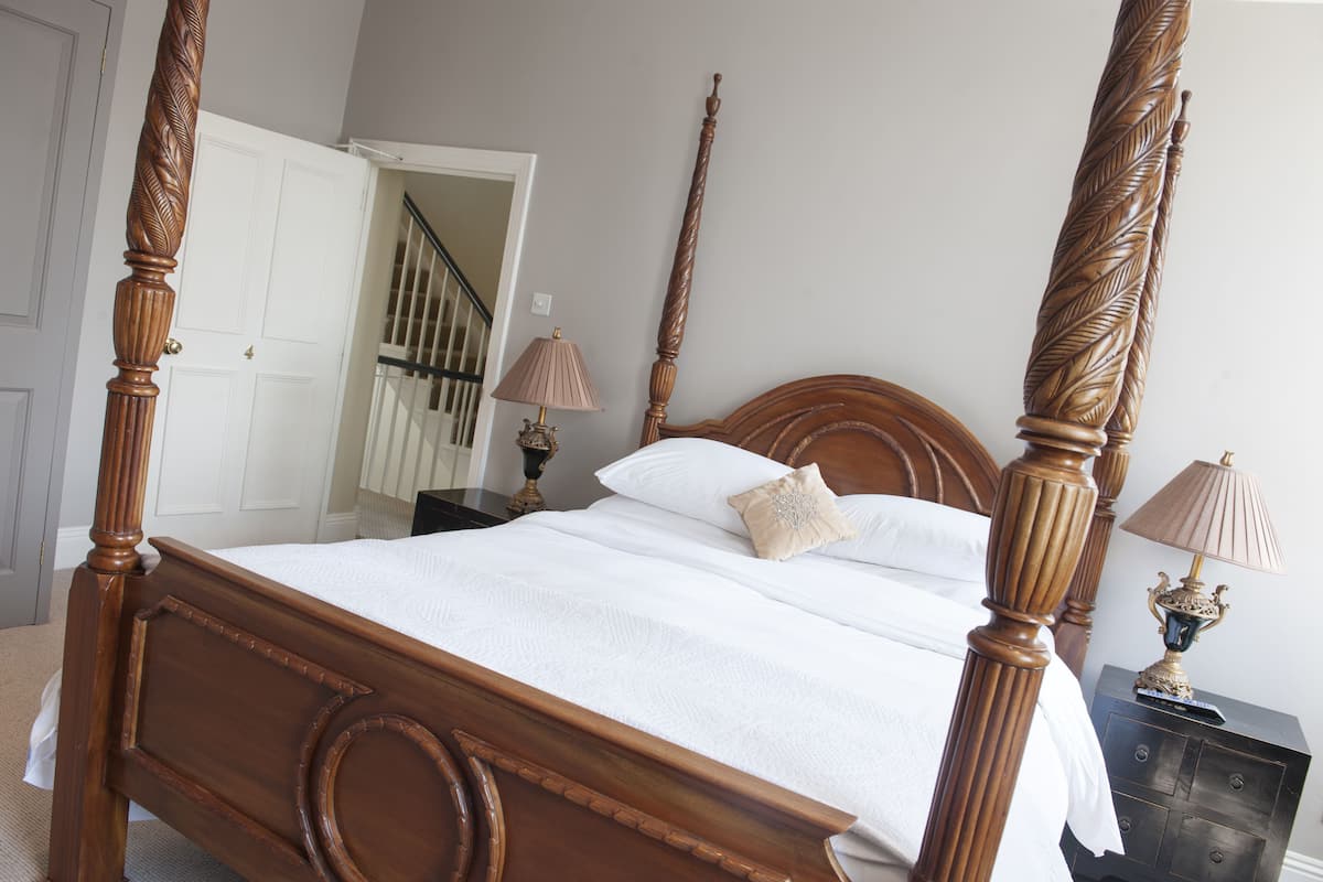 Un lit à baldaquin avec une tête de lit en bois, une table de chevet avec une lampe et une porte donnant sur un escalier.