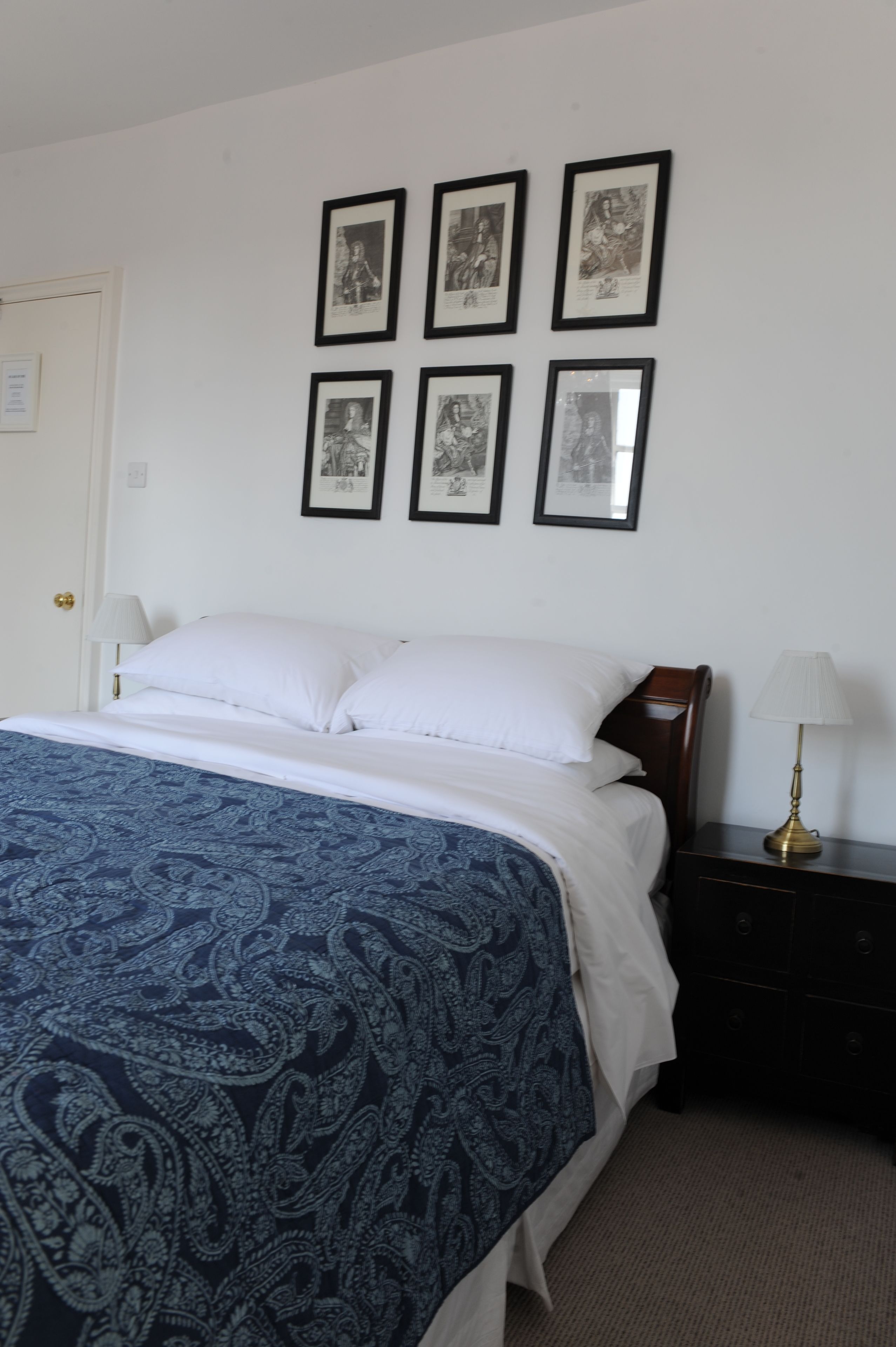 Un dormitorio con una cama, almohadas blancas, una colcha de tonos azules y dibujos, una mesita de noche con una lámpara, y seis cuadros en la pared.
