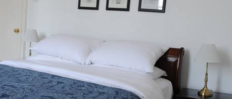 Un dormitorio con una cama, almohadas blancas, una colcha de tonos azules y dibujos, una mesita de noche con una lámpara, y seis cuadros en la pared.
