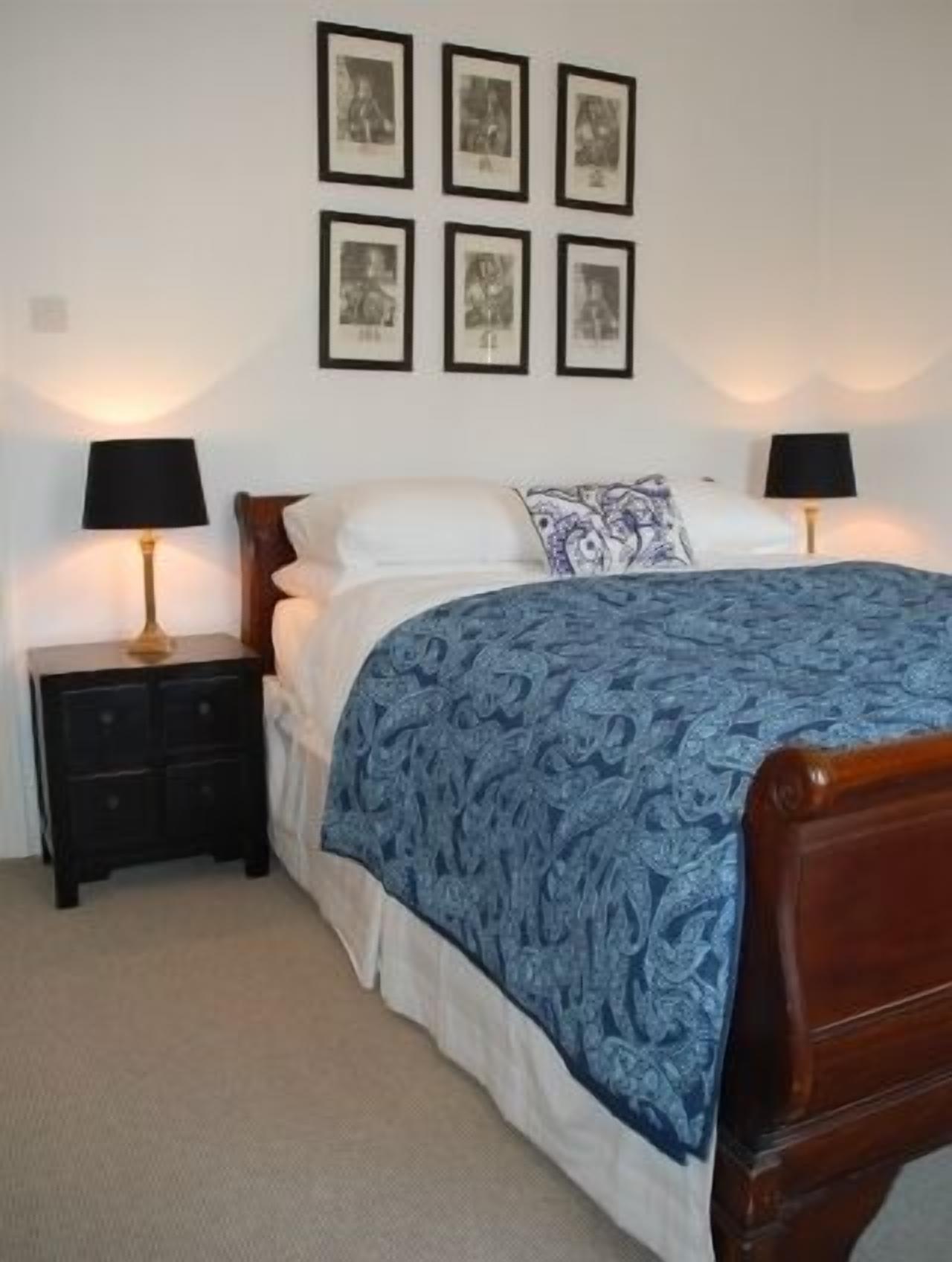 Un dormitorio con una cama de madera, dos mesitas de noche con lámparas, una colcha estampada y seis cuadros enmarcados en la pared.