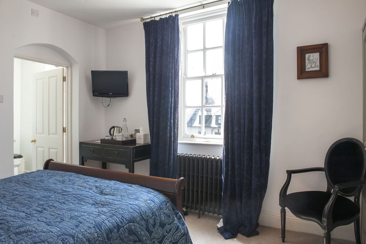 Une chambre à coucher comprenant un lit, une commode, une chaise, un téléviseur et une fenêtre avec des rideaux.