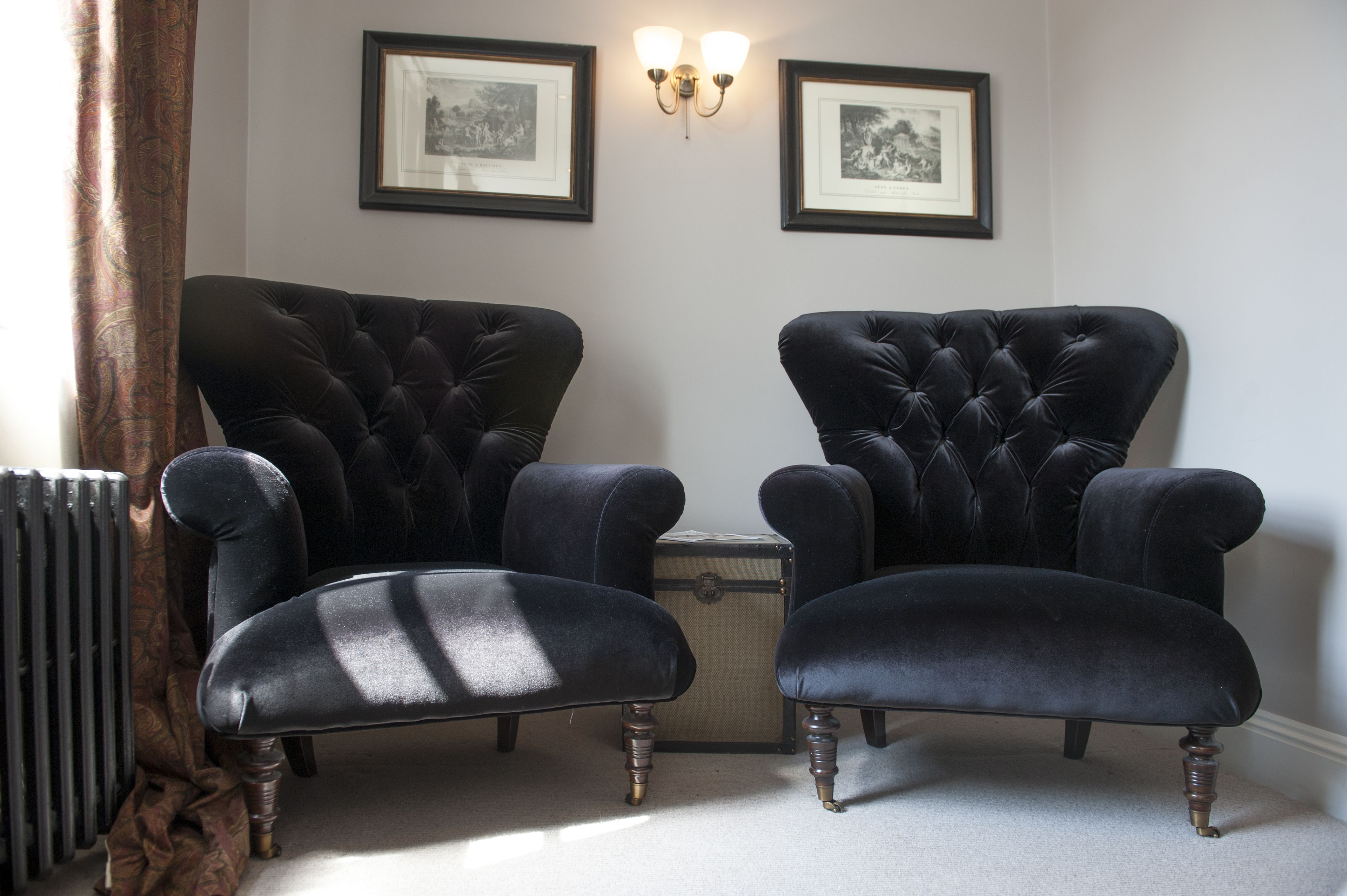 Two black upholstered armchairs with a classic design, positioned on a carpeted floor with a radiator and a small wooden chest of drawers between them.