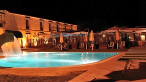 Uma área de piscina à noite com uma fonte, rodeada por um edifício com várias varandas e espaço ao ar livre.
