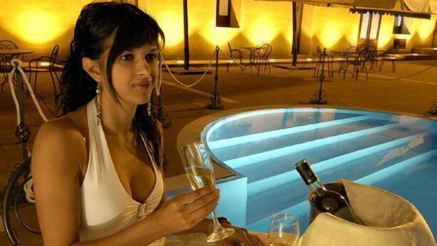 Uma mulher jantando à mesa perto de uma piscina, com velas, vinho e comida.