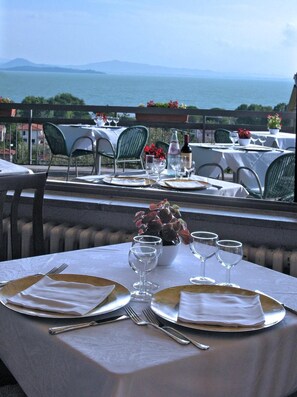 An outdoor dining area with tables set for two, overlooking a body of water and distant mountains.