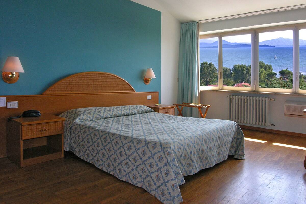 A bedroom with a bed, bedside tables, a lamp, a window with a view of the sea, and a radiator.